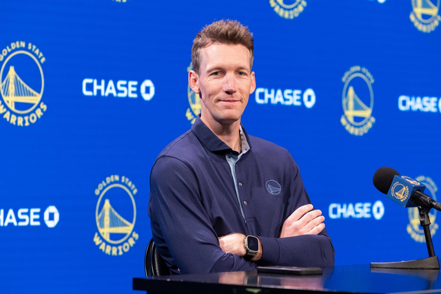 A person in a polo shirt stands with folded arms in front of a blue background adorned with Golden State Warriors logos and “CHASE” text. A microphone is nearby.